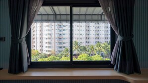 A room with a large window, curtains, and a view of a high-rise building with balconies and palm trees. - Phunaya Phuket Oldtown Hotel - SELF CHECK IN (Phuket)