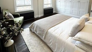 Une chambre avec un grand lit, deux fenêtres équipées de stores, un radiateur, une armoire et un fauteuil vert.