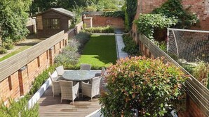 Un jardin avec une terrasse en bois, une petite table entourée de chaises et un bâtiment en briques doté d’une cheminée.