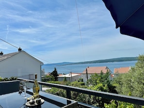 A balcony with a table, a bottle, and a glass. The view includes a body of water and distant hills. - Apartman Queen (Dramalj)