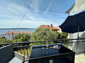 A balcony with a glass table, two chairs, and an umbrella. The view includes a body of water and buildings in the distance. - Apartman Queen (Dramalj)