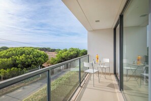 Ein Balkon mit Glasgeländer, einem Tisch mit Stühlen und Blick auf Grünflächen und eine Straße.