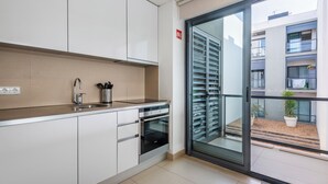 Una cocina moderna con armarios blancos, un lavamanos de acero inoxidable y un horno. Hay una puerta corrediza de vidrio que da a un balcón con vista a un edificio y un pequeño jardín.