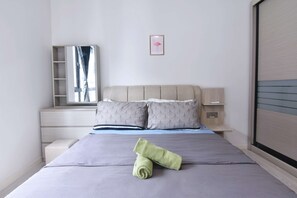 A neatly made bed with a folded towel, a bedside table with a lamp, a wall-mounted shelf, and a framed picture.