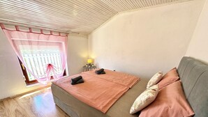 Um quarto aconchegante com piso de madeira, um sofá com manta rosa e almofadas, uma mesinha com abajur e uma janela com cortina.