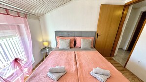 Uma cama de casal bem arrumada com roupa de cama rosa, uma mesa de cabeceira com abajur e uma janela com cortinas rosa.