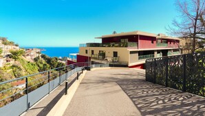 A modern building with a red and beige facade, situated on a hillside overlooking a coastal area.