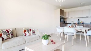 A modern living room with a sofa, a dining table with chairs, a kitchen area, and a pendant light.