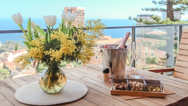 A wooden table with a vase of flowers, a champagne bottle, glasses, and a tray of nuts on a balcony with a view of buildings and the sea.