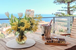 Une table en bois ornée d’un vase de fleurs, d’une bouteille de champagne, de verres et d’un plateau de noix, le tout placé sur un balcon offrant une vue sur les bâtiments et la mer.