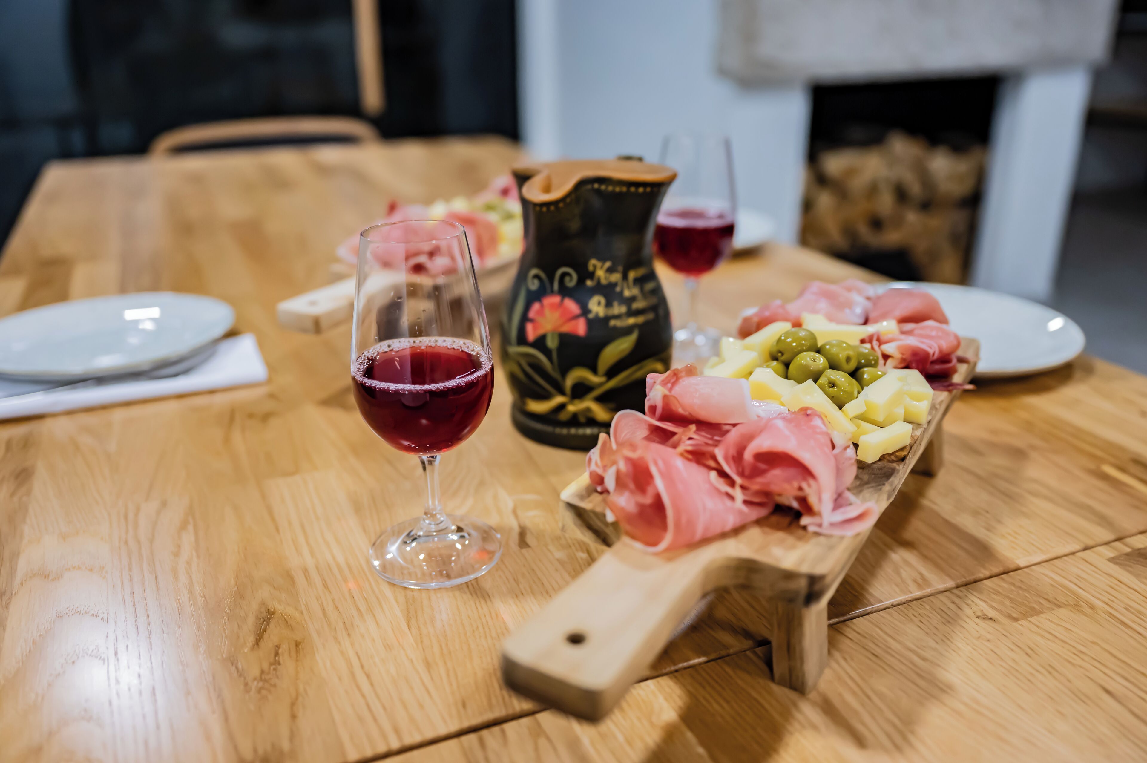 Une table en bois, sur laquelle on a disposé une planche de charcuterie, un verre de vin rouge et un pichet décoratif.