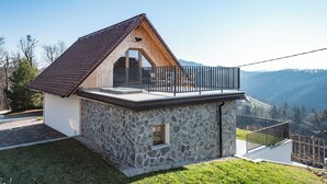 Una casa moderna con fachada de piedra, un balcón con barandilla metálica y vistas a las montañas circundantes.