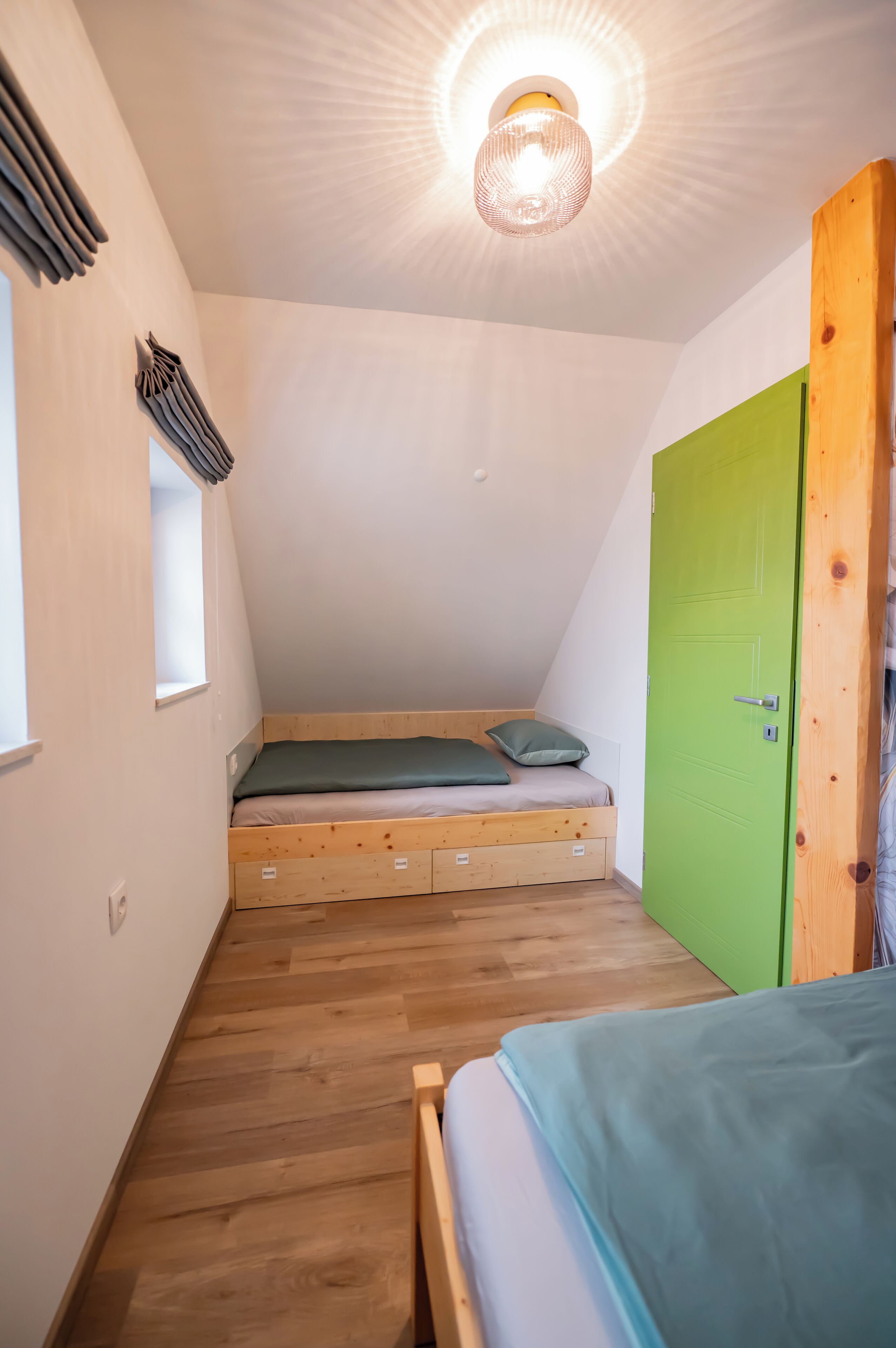 A small, triangular attic bedroom with a sloped ceiling, a single bed, a green door, and a small window.