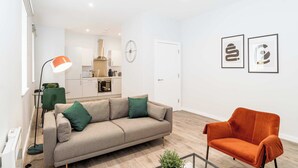 Ein modernes Wohnzimmer mit einer Couch, einem orangefarbenen Sessel und einer Stehlampe. Im Hintergrund ist eine Küche mit weißen Schränken und einer Uhr an der Wand zu sehen.