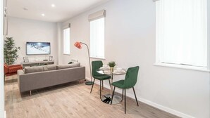 Ein modernes Wohnzimmer mit grauem Sofa, einem runden Glastisch, zwei grünen Stühlen, einem an der Wand befestigten Fernseher und einem großen Fenster mit Jalousien.
