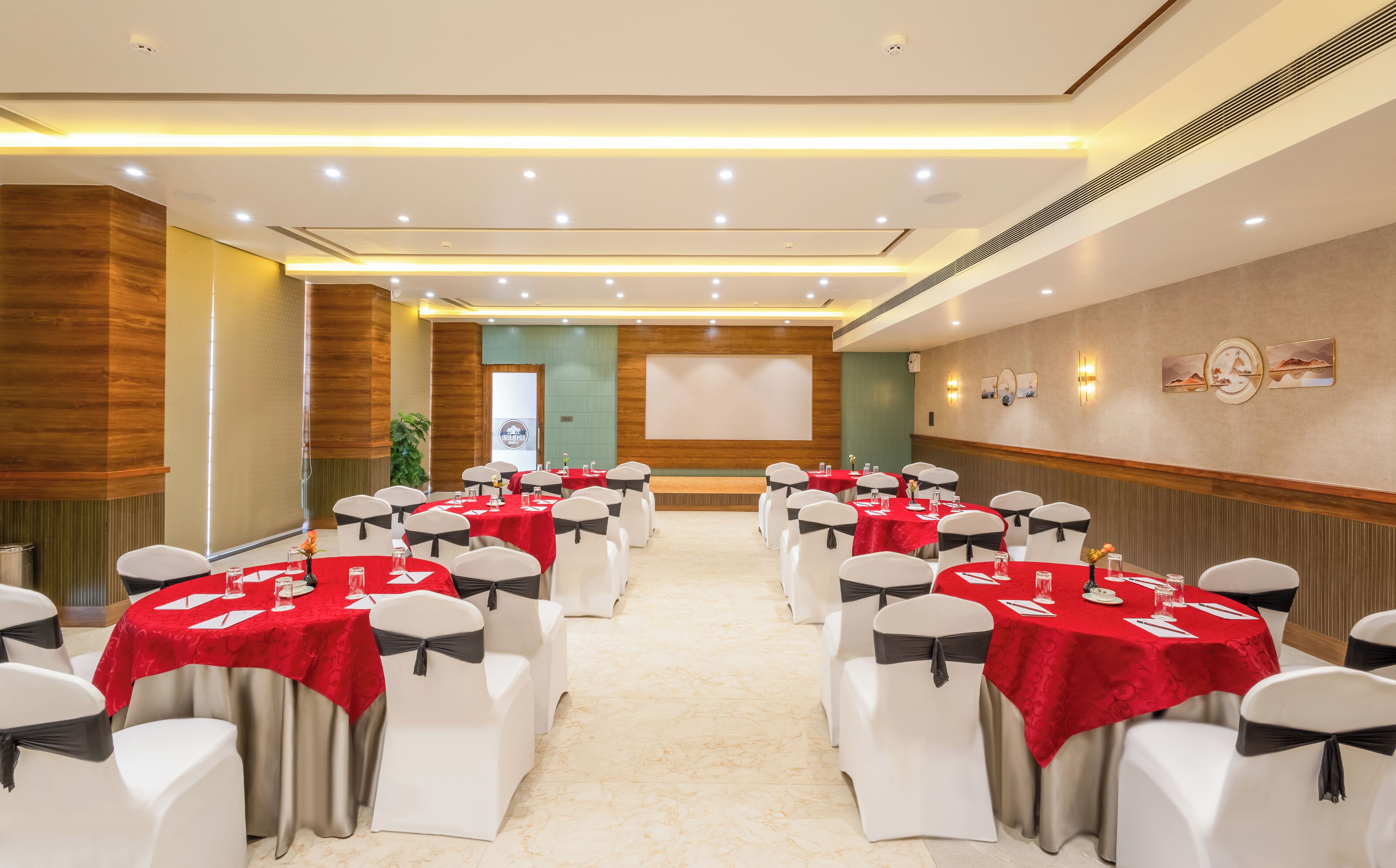 A conference room with rows of white chairs and red tablecloths, equipped with ceiling lights and a large screen at the front.