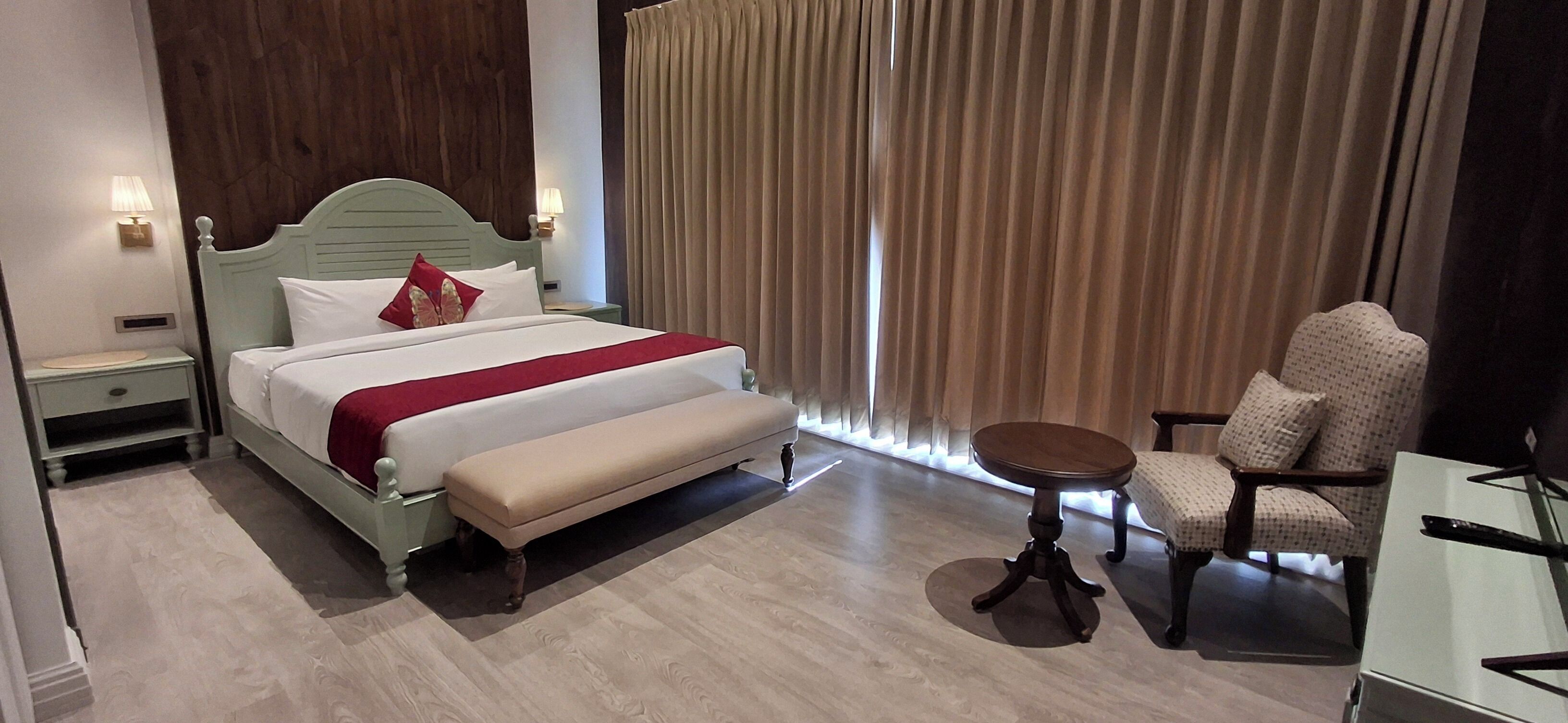 A hotel room with a bed, a bench, a chair, a small table, and a window with curtains.