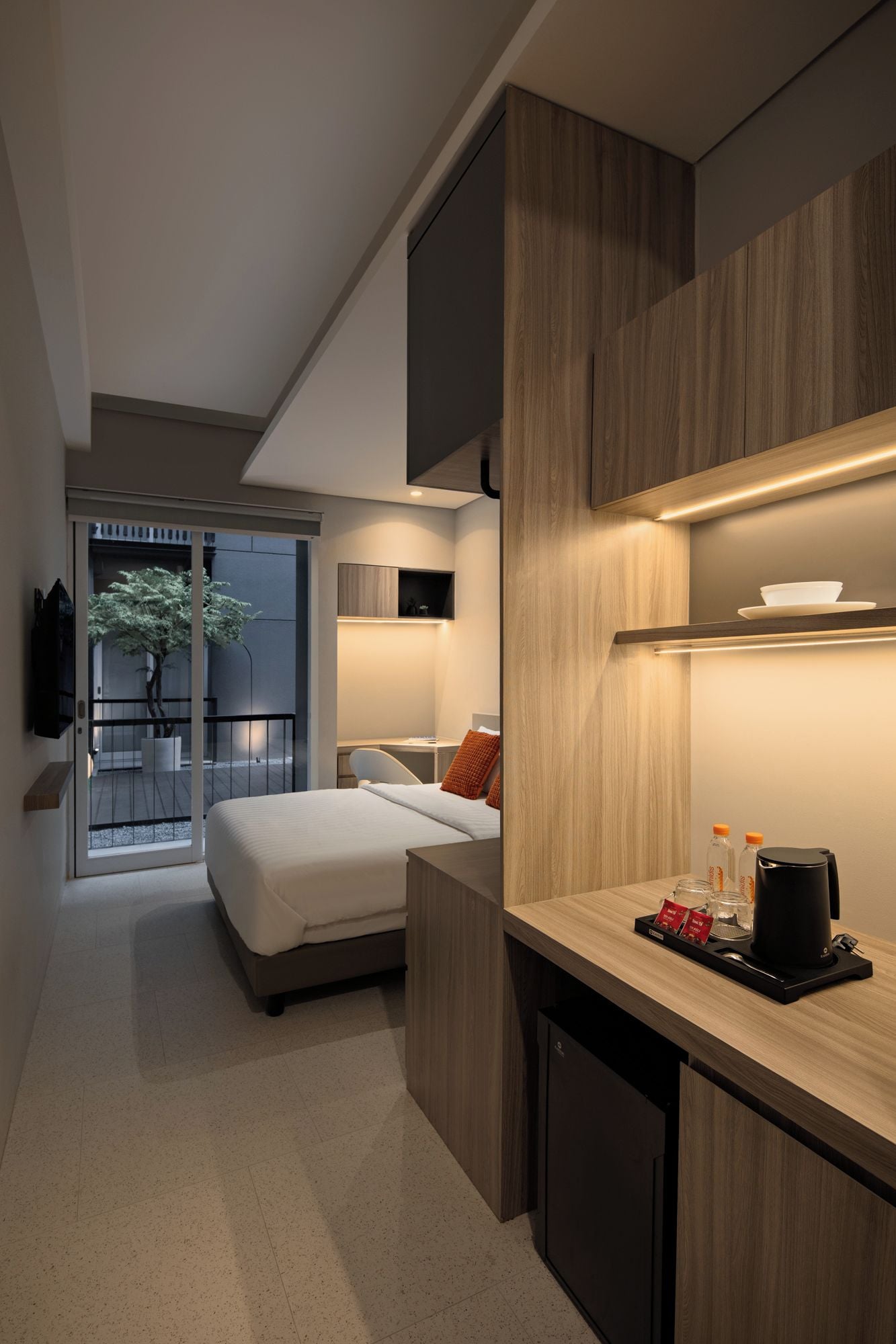 Una habitación de hotel moderna con cama, escritorio, una kitchenette pequeña y un televisor montado en la pared.