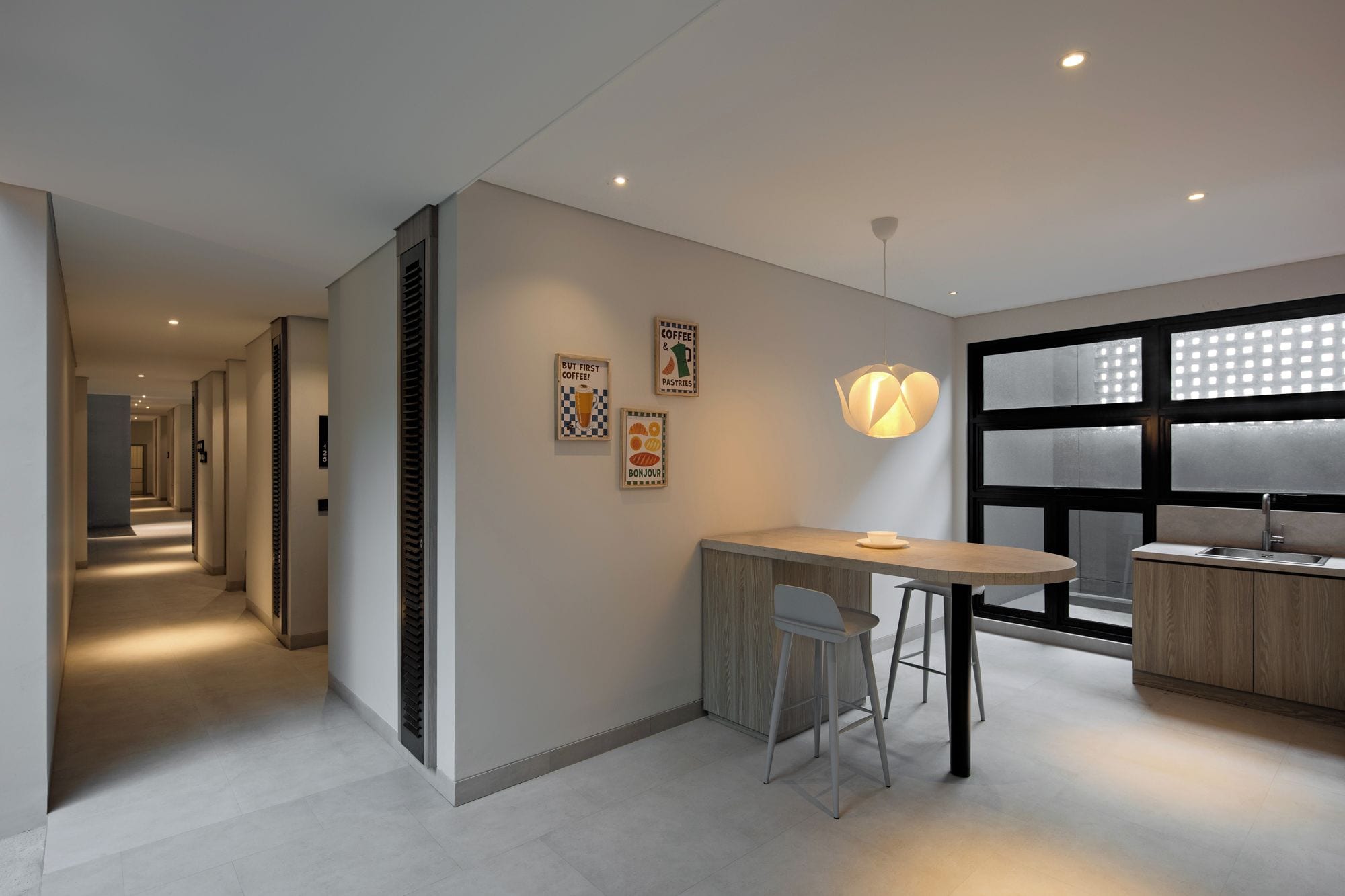 A modern hotel room with a kitchenette, a dining area with a table and chairs, and a large window with blinds.