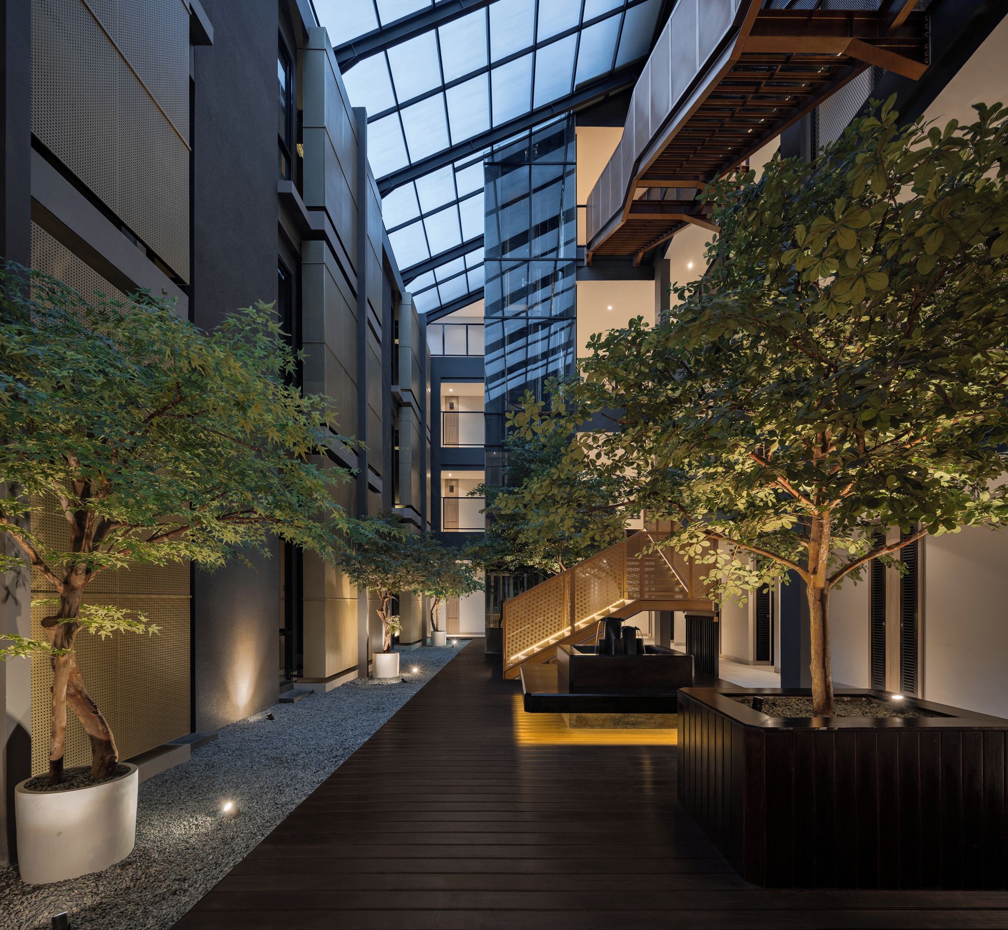 A modern atrium with a wooden walkway, glass ceiling, and indoor trees.