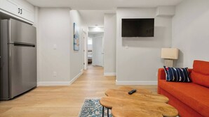 A modern apartment with a kitchen, a living area with a sofa and a TV, and a hallway leading to a bedroom. - 1BR Lower Level Suite with Secure Entry & Laundry (Calgary)