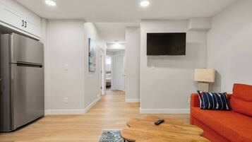 A modern apartment with a kitchen, a living area with a sofa and a TV, and a hallway leading to a bedroom.