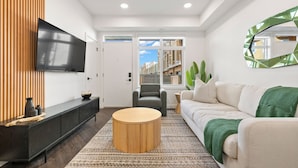A modern living room with a flat-screen TV, a sofa, a round coffee table, and a large window with a view of buildings. - Designer-Inspired Stay 1BR + Sauna/Massage Chair (Calgary)