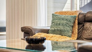 A leather sofa with a textured throw blanket and a cushion, a glass coffee table with a bowl, and a view of a building through a window with curtains. - Bright Nature Escape 2BR Walkout Suite w/ Patio (Calgary)