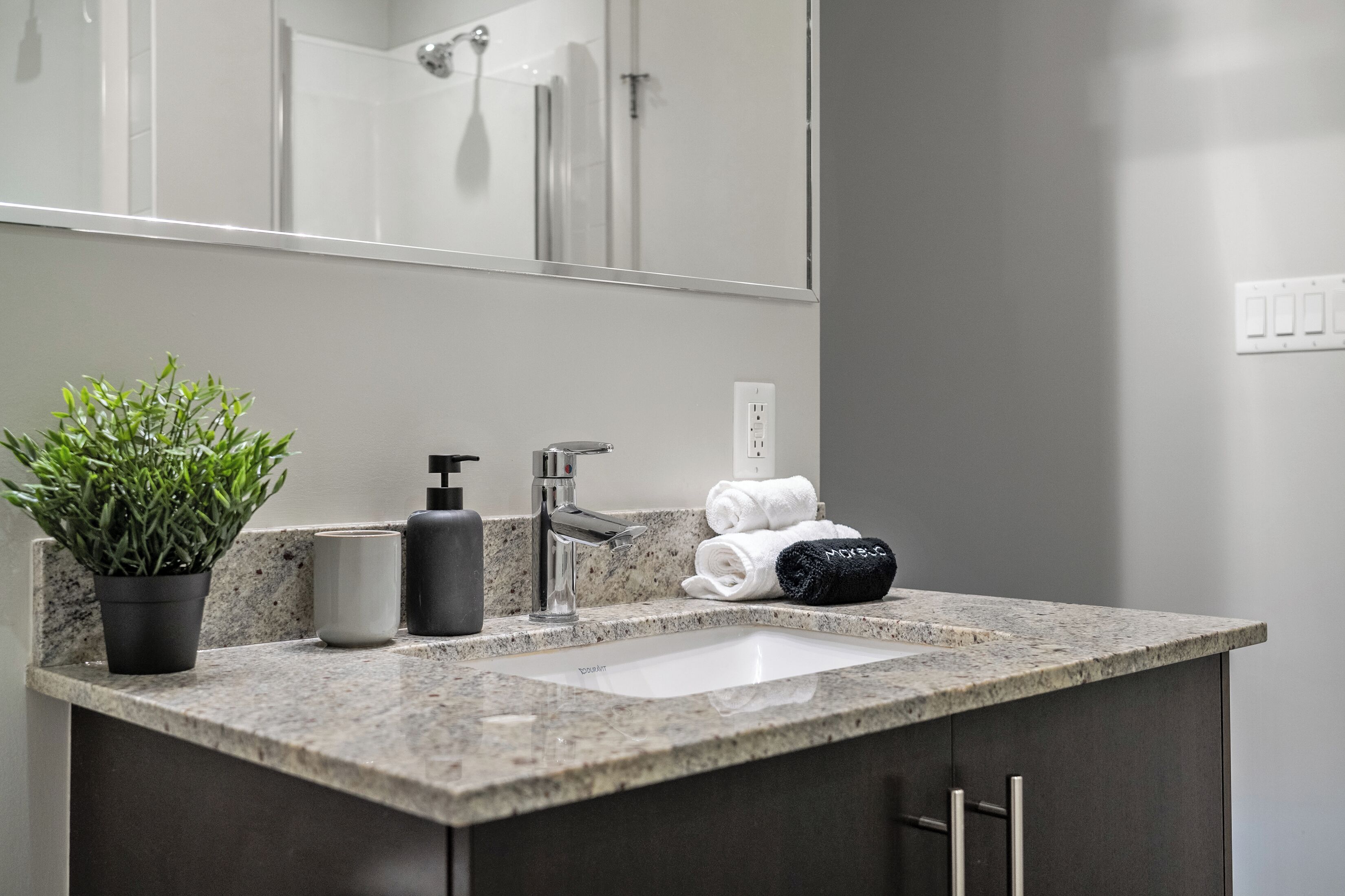 A bathroom with a granite countertop, a sink, a mirror, a potted plant, a soap dispenser, and towels.