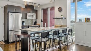 A modern kitchen with a central island, bar stools, stainless steel appliances, and a large window offering a city view.