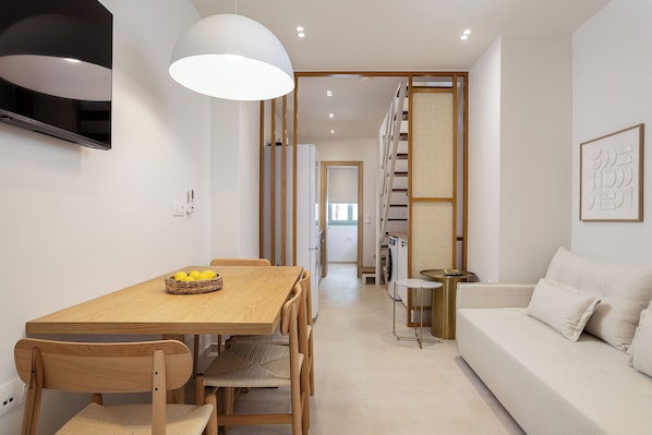 A modern interior with a wooden dining table, chairs, a sofa, a TV, and a kitchen area.