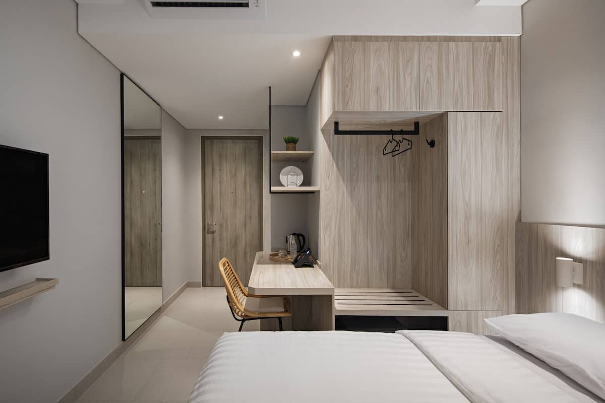 A modern hotel room with a large flat-screen TV, a wooden desk with a chair, a bed, and a small seating area with a chair and a table.