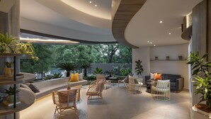 Ein modernes Interieur mit gewölbter Decke, einer Sitzecke mit Korbstühlen, einem Sofa und einem Esstisch. Übertöpfe mit Pflanzen und Blick auf Bäume draußen.