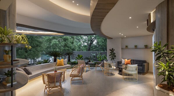 A modern interior with a curved ceiling, a seating area with wicker chairs, a sofa, and a dining table. There are potted plants and a view of trees outside. - FULMAR Dago (Bandung)