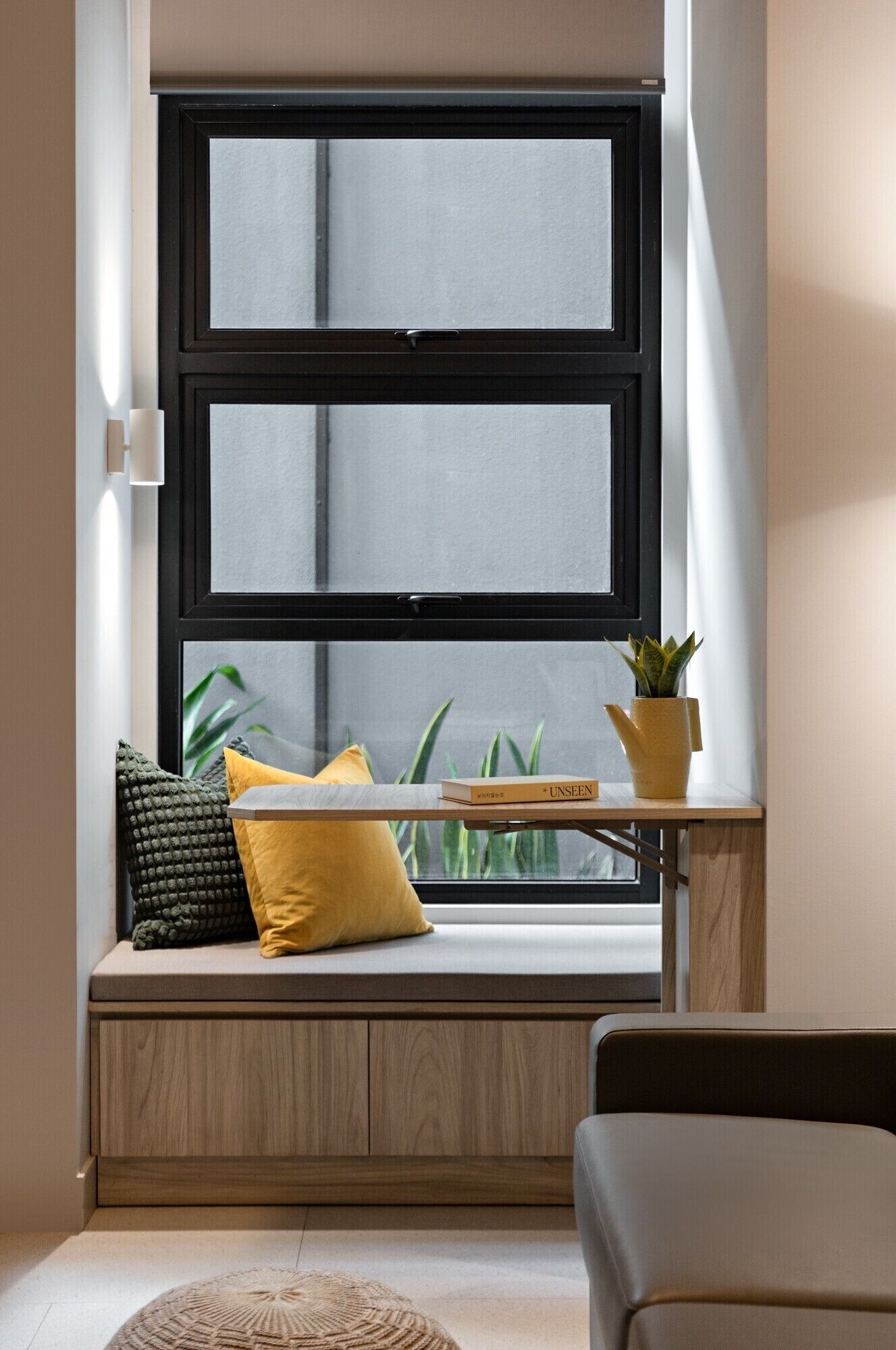 A room with a built-in bench under a large window, a small table with a book, and a potted plant.