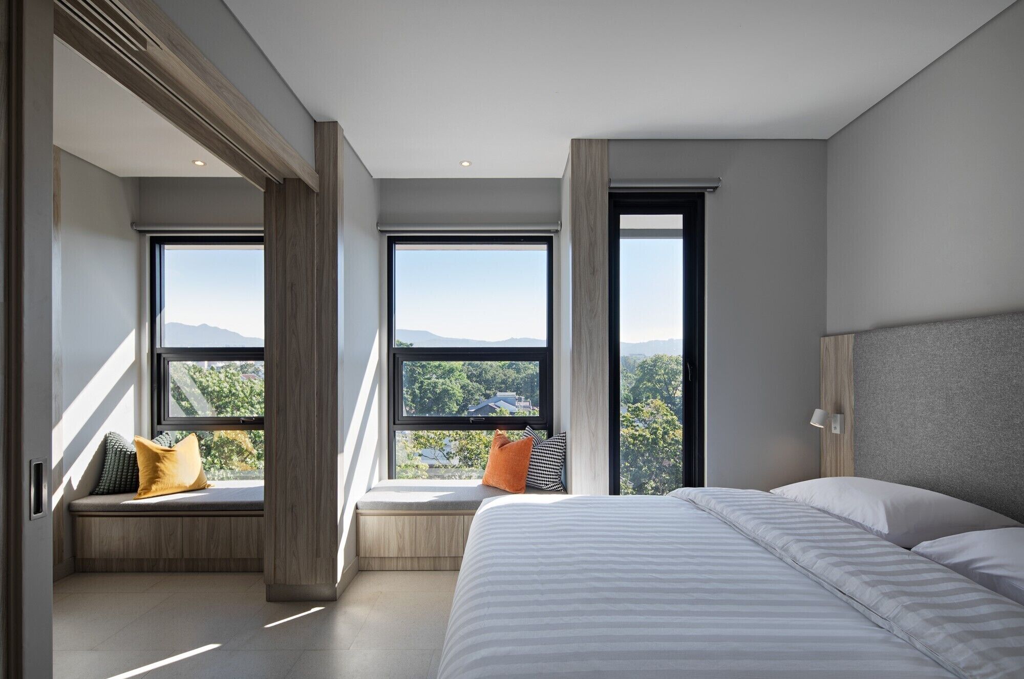 A modern bedroom with a large bed, built-in bench with cushions, and large windows offering a mountain view.