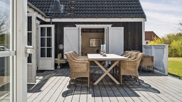 Een houten terras met een tafel en rieten stoelen, een witte deur en een donker betegeld dak.
