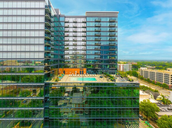 A modern high-rise building with multiple balconies, a rooftop pool, and a glass facade reflecting the surrounding cityscape.