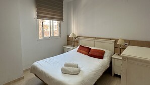 Une chambre à coucher comprenant un lit, une table de chevet, une lampe, une fenêtre avec des stores et un luminaire au plafond.
