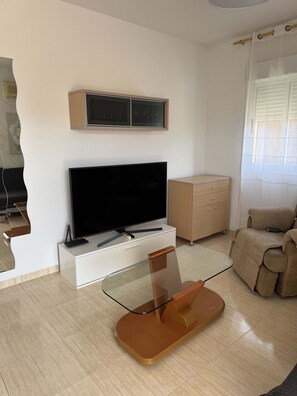 A living room with a flat-screen TV, a glass coffee table, a beige sofa, and a wooden cabinet.