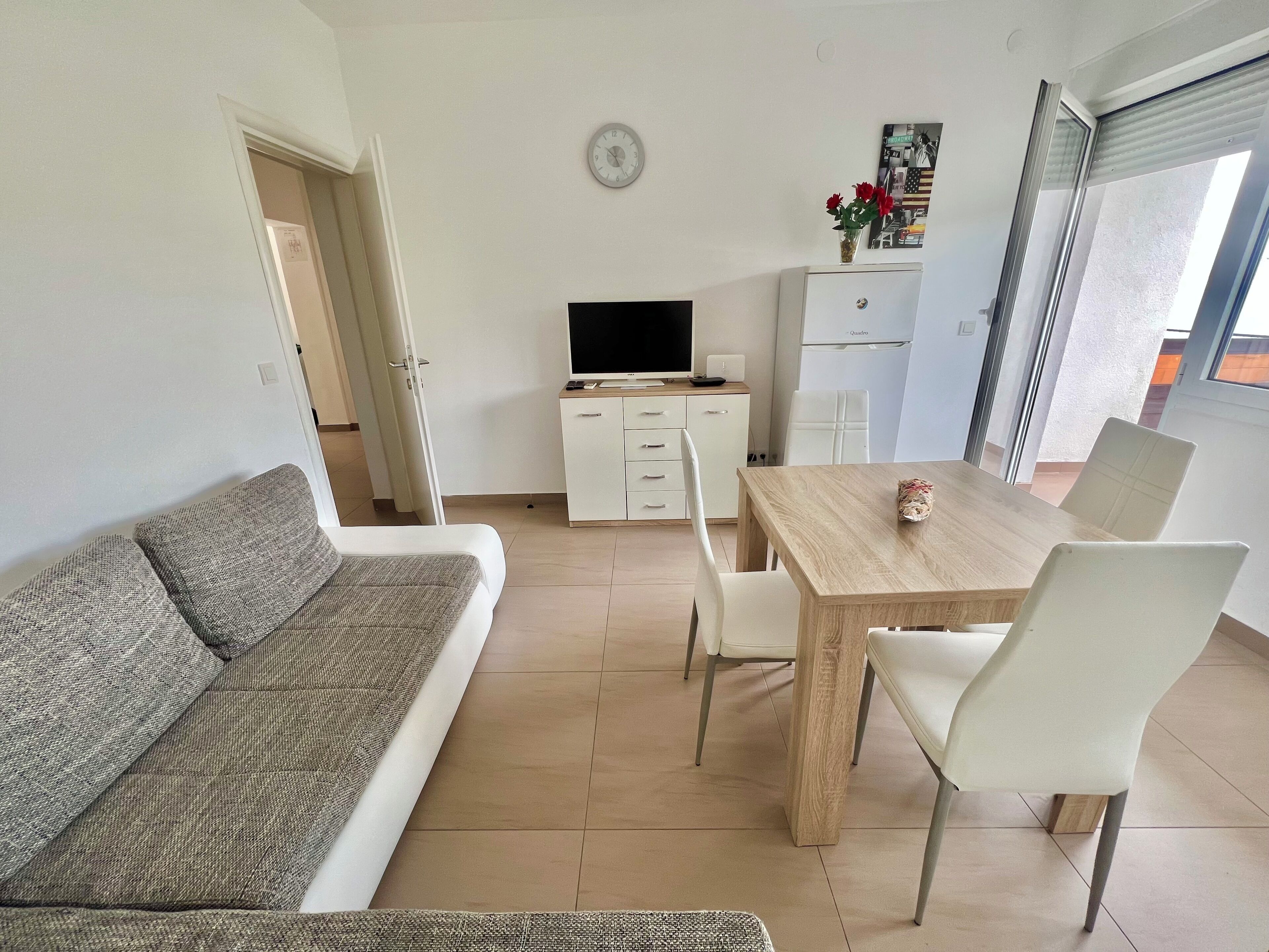 Una sala de estar moderna con un sofá, una mesa de comedor con sillas, un televisor, un refrigerador y un reloj en la pared.