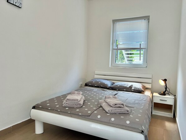 A neatly made bed with a polka dot bedspread and pillows, a bedside table with a lamp, and a window with blinds. - Apartman Velly  (Jadranovo)