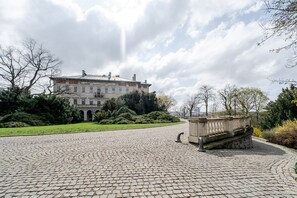 Une cour pavée de pierres, avec une fontaine, un grand bâtiment en arrière-plan et une pelouse bien entretenue.