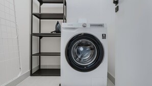 A compact, white front-loading washing machine in a modern laundry room with a tiled floor and metal shelving unit. - Blueground | Prague 3, close to offices & mall (PRG-20) (Prague)