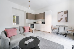 Un apartamento moderno con un sofá gris, una mesa de centro redonda, una cocina con armarios blancos y un comedor con una mesa pequeña y sillas.
