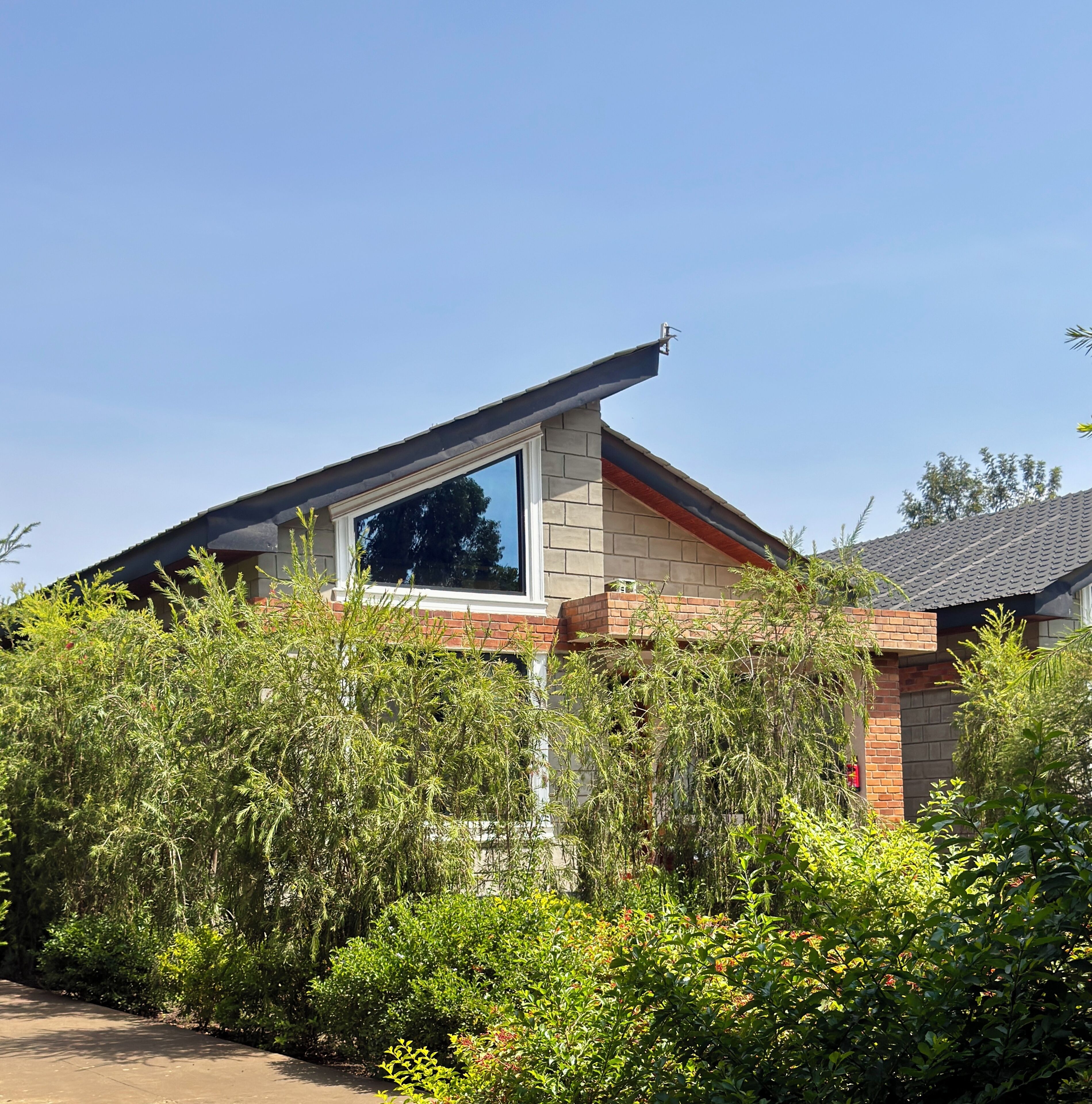 A house with a large window, brick walls, and dense greenery in front.