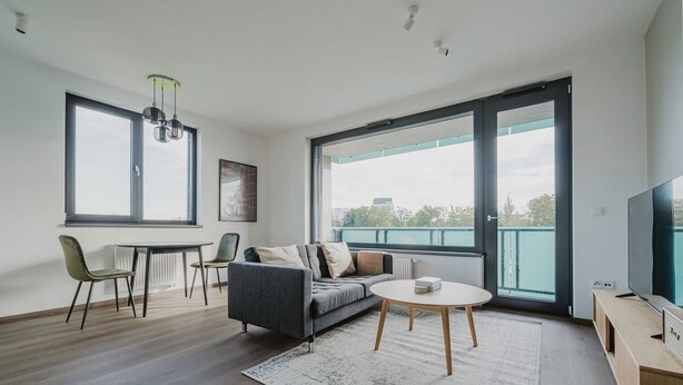 Ein modernes Wohnzimmer mit grauem Sofa, rundem Couchtisch und Fernseher auf einem Holzständer. Zwei Sessel, ein Esstisch und ein großes Fenster mit Blick auf die Stadt sind ebenfalls vorhanden.