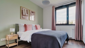 Ein Schlafzimmer mit Bett, Nachttisch mit Lampe, Fenster mit Vorhängen und Heizkörper.