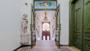 Un corridoio con una statua, un soffitto decorato e un pavimento a motivi che porta a una porta con un cartello.