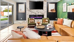 A modern living room with a fireplace, green and brown upholstered chairs, a leather sofa, and a flat-screen TV mounted above the fireplace. - The Ridge: Family | Pickleball | Pool | Hot Tub (Phoenix)